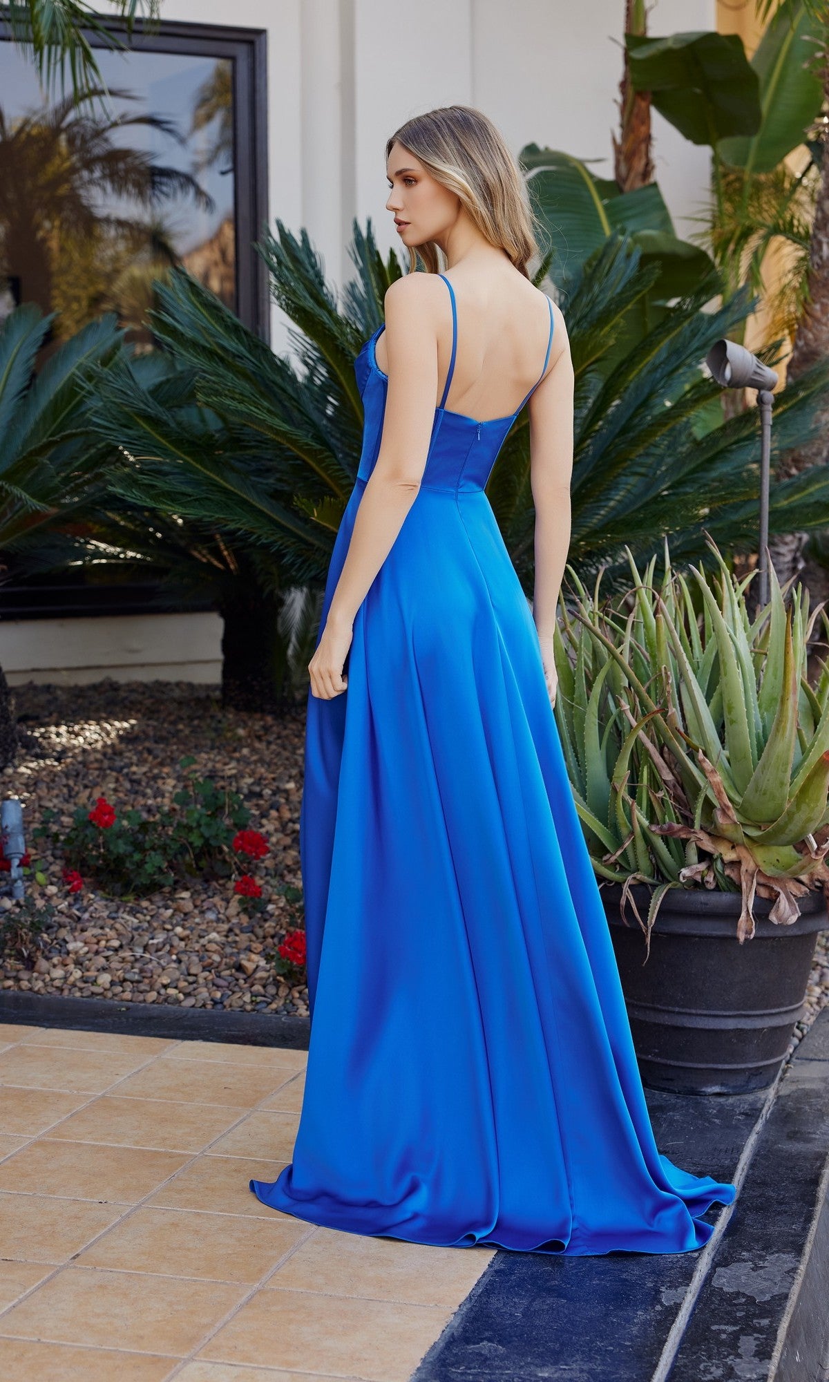 A woman in the Nox Anabel A1744 long prom dress stands outdoors on a tile walkway, facing away, near green plants and a garden bed with rocks and flowers.
